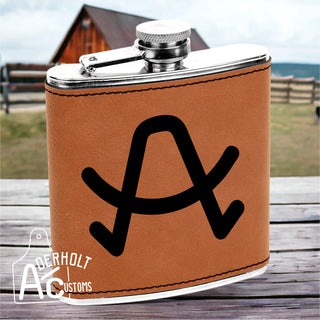 Brown leather flask with black livestock brand engraved on it, sitting on a wooden surface, with a blurred background of a barn and fence.