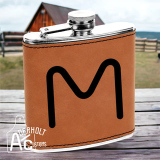 Brown leather flask with black livestock brand engraved on it, sitting on a wooden surface, with a blurred background of a barn and fence.